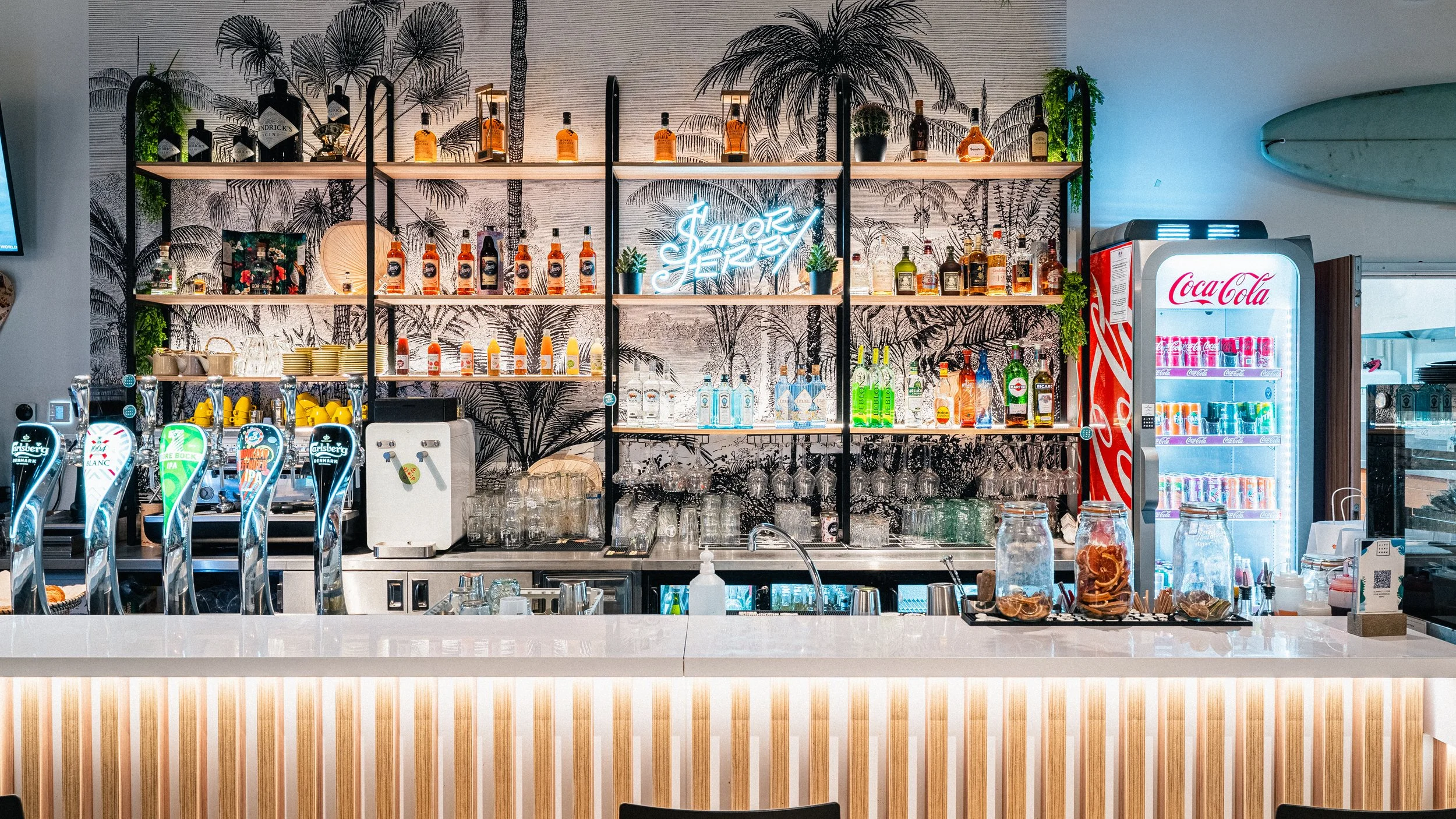 Bar avec étagères de bouteilles de boissons alcoolisées, un distributeur de boissons et un réfrigérateur Coca-Cola, décor tropical avec un mur de palmier en arrière-plan, néon « Saint-Tropez »