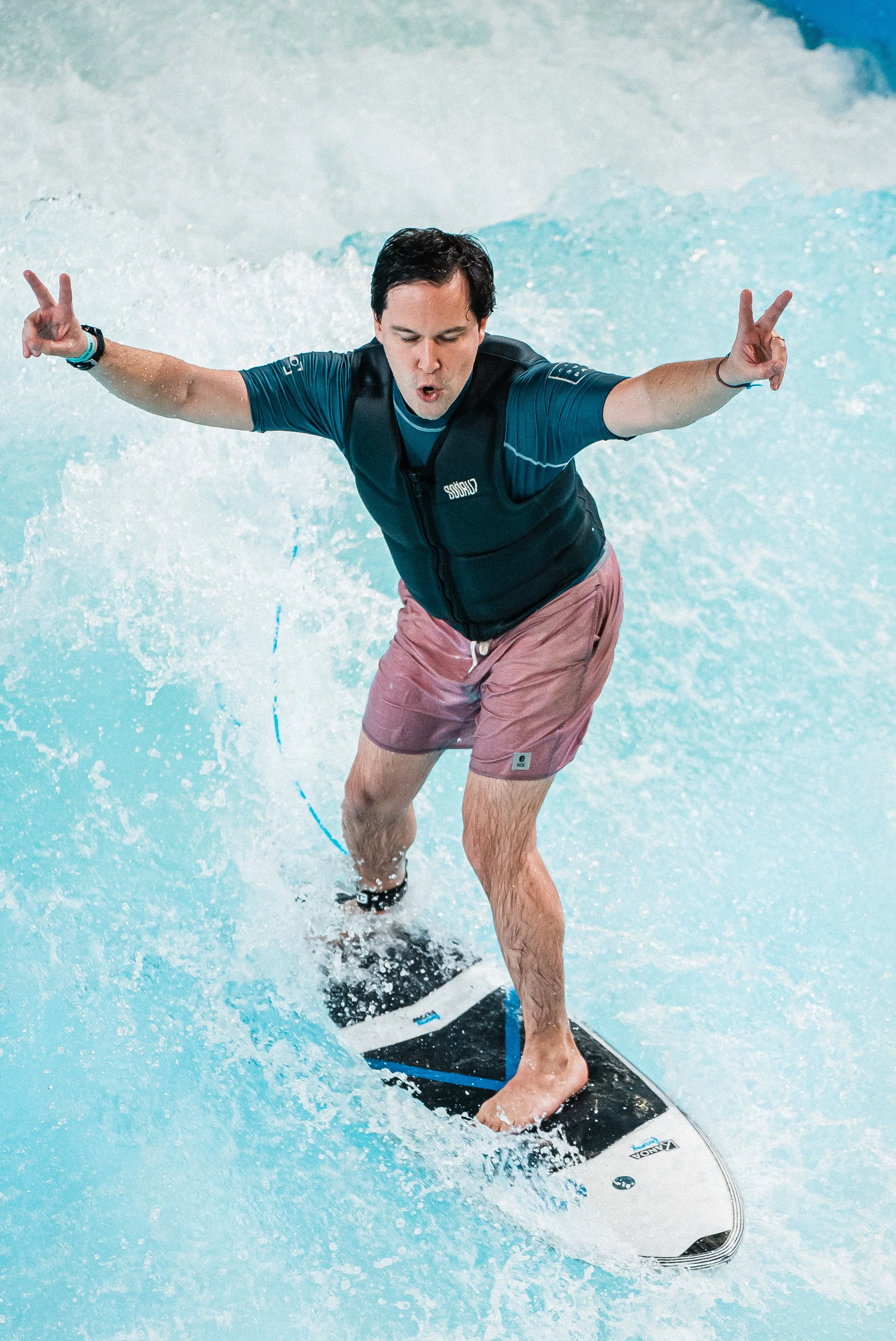 Un homme en maillot de bain et veste, debout sur une planche de surf dans une piscine, faisant un geste de paix avec les mains